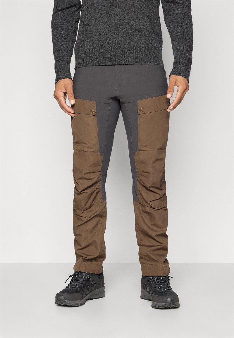 Man in een donkergrijze en bruine cargo broek met knoopzakken op de bovenbenen en zwarte wandelschoenen tegen een effen achtergrond.