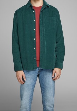 Muž v zelené manšestrové košili přes červené tričko s kulatým výstřihem, v kombinaci se světlými modrými džínami, stojící před jednobarevným šedým pozadím.