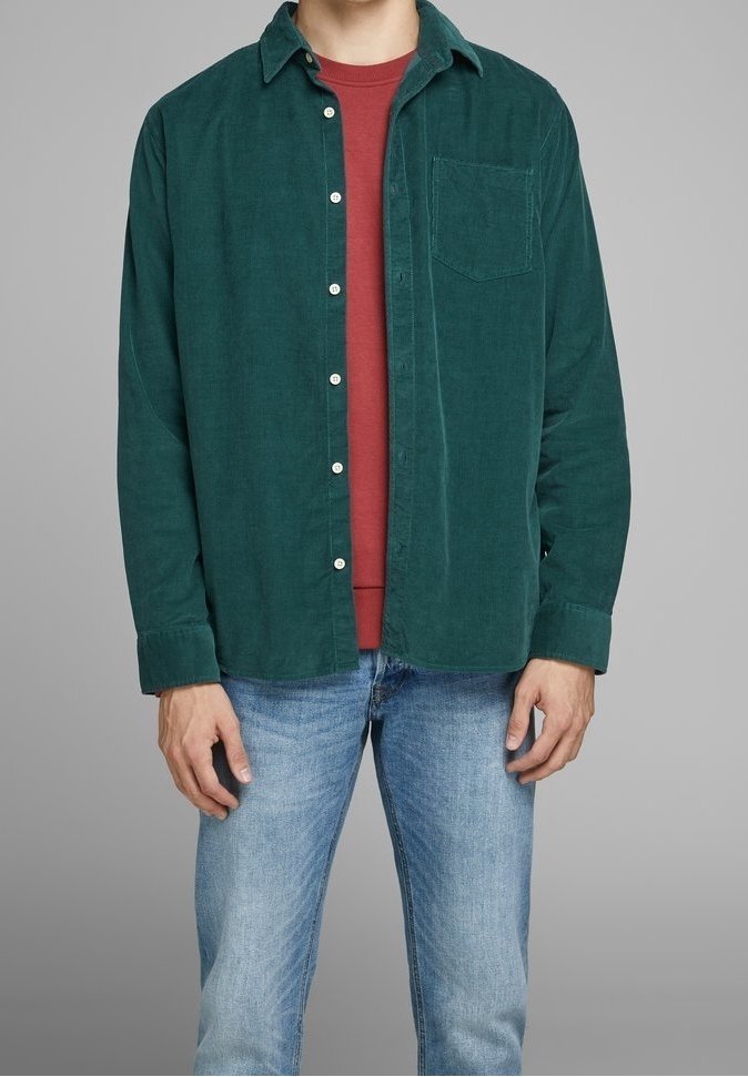 Homme portant une chemise en velours côtelé verte à boutons par-dessus un t-shirt rouge à col rond, associé à un jean bleu clair, debout devant un fond gris uni.