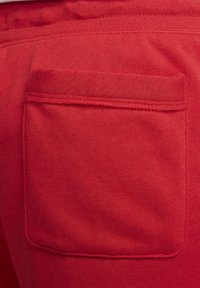 Gros plan d'une poche arrière cousue sur un pantalon en tissu rouge avec des coutures visibles et un rabat en tissu en haut.