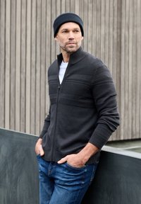 Suéter gris oscuro con cremallera y patrón acanalado, combinado con un gorro negro y jeans azules. Textura suave y diseño ajustado.