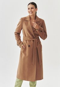 Abrigo de lana marrón con diseño de doble botonadura, amplios solapas, cinturón autoadherente y dos bolsillos frontales. Textura suave, estilo hasta la rodilla.