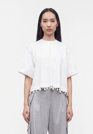 Woman with long black hair wearing a white short-sleeve top with pearl trim and loose gray pants, standing against a plain background.