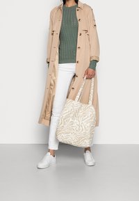 Femme portant un trench beige, un pull en maille vert, un pantalon blanc, des baskets blanches, tenant un sac fourre-tout à motifs beige et blanc.
