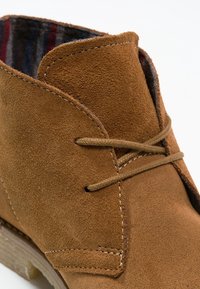 Bruine suede enkellaars met een ronde neus, zijnaadstiksels en twee veters. De binnenvoering heeft een gestreepte stof in rood en blauw.