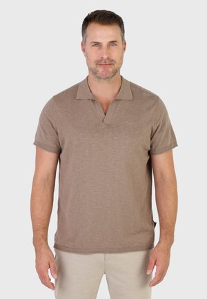 Mężczyzna z krótkimi ciemnymi włosami i jasnym zarostem, ubrany w taupe krótkorękawową koszulkę polo i beżowe spodnie, stojący przodem na jednolitym szarym tle.