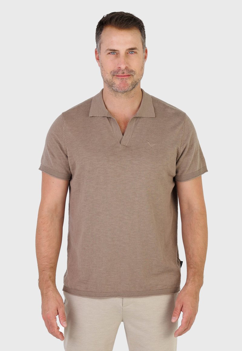 Homme aux cheveux courts et foncés avec une barbe légère, portant un polo à manches courtes couleur taupe et un pantalon beige, face à l'avant sur un fond gris uni.