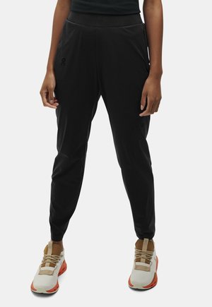 Personne portant un pantalon noir fuselé avec une ceinture élastique et des baskets blanches avec des touches orange et beige, debout devant un fond uni.