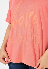 Camisa de color coral con texto texturizado brillante "Love Out Loud", llevada por una persona que sostiene el dobladillo de la camisa, combinada con jeans azul oscuro.