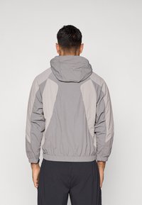 Pelēks kapucejājs džemperis ar gaismas un tumšā pelēka paneļu kombināciju, elastīgām aprocēm un sašaurinātu jostasvietu, izgatavots no viegla materiāla.