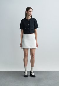 Žena oblečená v čiernej krátkej saku s gombíkmi, bielej textúrovanej mini sukni, béžových ponožkách a čiernych lodičkách Mary Jane stojí pri šedej stene.