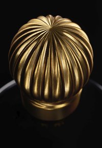 Złoty pokrętło o żeberkowanej teksturze i zaokrąglonym wierzchołku. Materiał wydaje się metaliczny o połyskującym wykończeniu, umieszczony na czarnym tle.