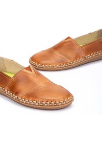 Zapatos de cuero color marrón claro con suela plana, que presentan detalles cosidos y un acento trenzado a lo largo del borde. El interior tiene un forro de color verde brillante.