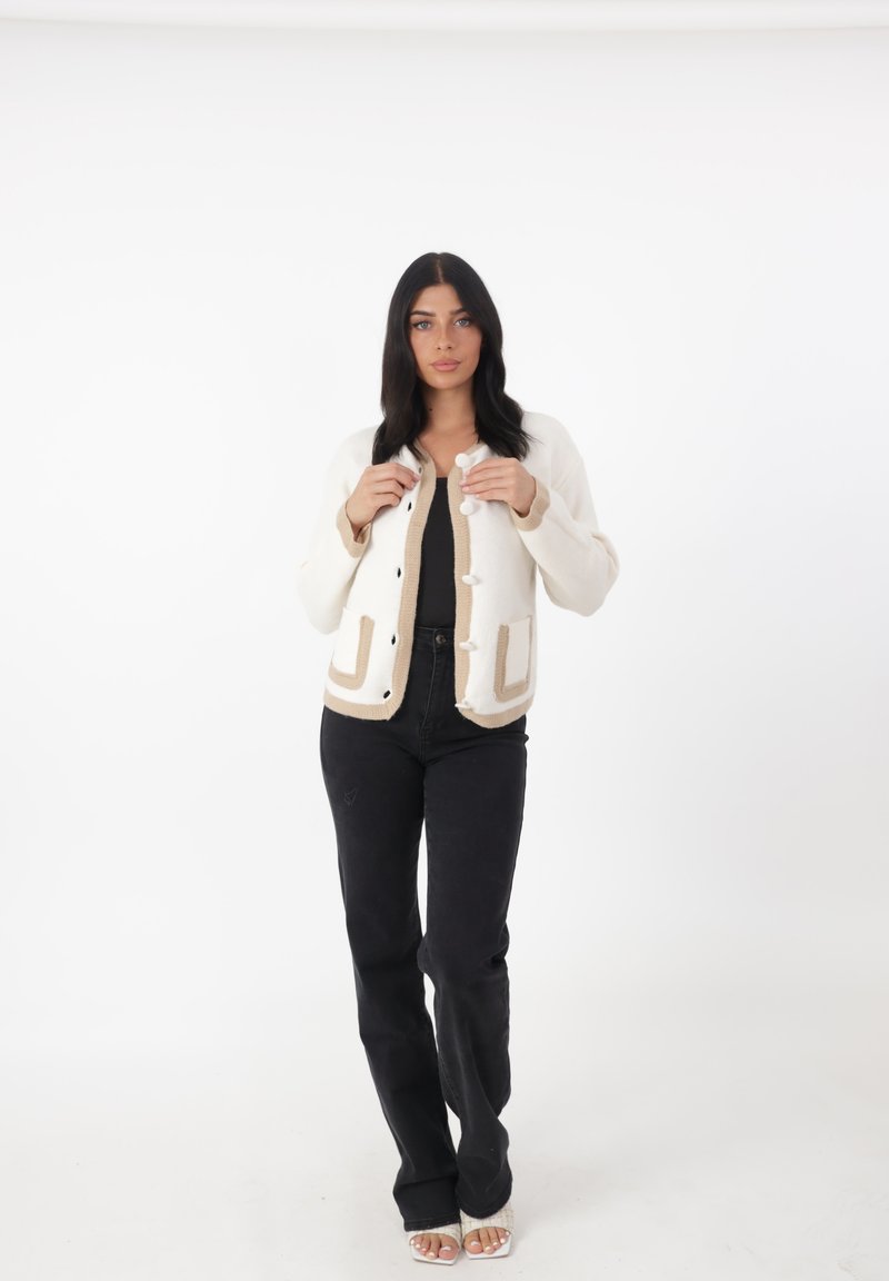 Cardigan crema con dettagli beige, adornato con bottoni rotondi. Abbinato a pantaloni neri, in piedi su uno sfondo semplice.
