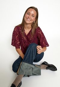 Camicia in pizzo borgogna con maniche corte, abbinata a jeans in denim rilassati. Scarpe nere con dettagli testurizzati. Posizione seduta su uno sfondo bianco.