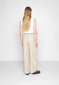 Kvinna med långt vågigt hår som bär en ärmlös vit topp, pastellfärgade blommiga vida byxor och svarta sandaler, stående mot en enfärgad bakgrund.