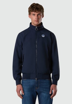 Giovane uomo di fronte che indossa una giacca blu navy con zip e un piccolo logo rotondo bianco e blu sul petto a sinistra, abbinata a jeans blu.