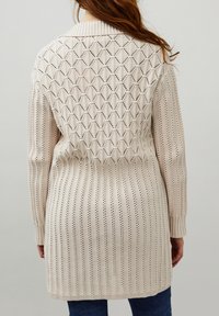 Gilet en tricot beige avec un motif texturé, présentant des motifs verticaux et des découpes en losange, des poignets côtelés et un col.