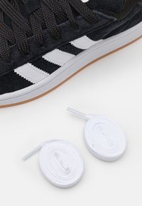 Baskets en daim noir avec des rayures en cuir blanc, des lacets tressés noirs et une semelle en caoutchouc blanc. Comprend deux lacets blancs roulés.