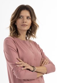 Cardigan rosa realizzato in morbido tessuto, caratterizzato da un collo rotondo, chiusura con bottoni e maniche lunghe con polsini a coste. Gli accessori includono braccialetti.