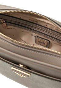 Intérieur de sac à main gris texturé avec fermeture éclair dorée, comprenant plusieurs compartiments et une étiquette logo "GUESS" sur une doublure en tissu beige.