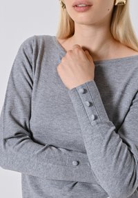 Maglione grigio a maglia con scollatura ampia. Presenta maniche lunghe e dettagli decorativi con bottoni sui polsini. Texture morbida e design aderente.