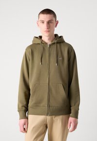 Olijfgroene rits-hoodie gemaakt van zachte stof, met een capuchon met trekkoord, voorzakken en ribgebreide manchetten. Geborduurd logo op de borst.
