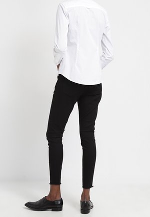 Osoba ubrana w dopasowaną białą koszulę z długim rękawem, czarne wąskie spodnie z surowymi wykończeniami u dołu oraz czarne skórzane mokasyny, stoi odwrócona tyłem.