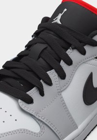 Κοντινό πλάνο ενός γκρι και λευκού αθλητικού παπουτσιού Nike Air Jordan με μαύρα κορδόνια, μαύρο λογότυπο Swoosh, κόκκινο περίγραμμα και το λογότυπο Jumpman στη γλώσσα.