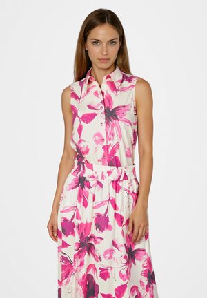 Junge Frau trägt ein ärmelloses weißes Kleid mit einem rosa und dunklen Blumenmuster, steht vor einem schlichten weißen Hintergrund.