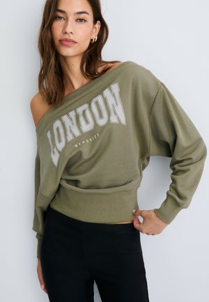 Junge Frau mit braunen Haaren trägt ein olivgrünes Schulterfreies Sweatshirt mit "LONDON MEMORIES"-Print und schwarze Hose und posiert vor einer weißen Wand.