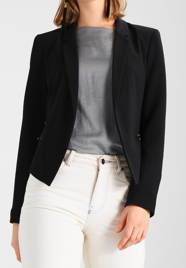 Blazer negro con un diseño ajustado, que presenta un cierre de un solo botón y dos bolsillos frontales, combinado con una blusa gris y pantalones blancos.