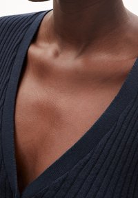 Detailní záběr na horní část hrudi a klíční kost osoby, která má na sobě tmavý žebrovaný výstřih do V.