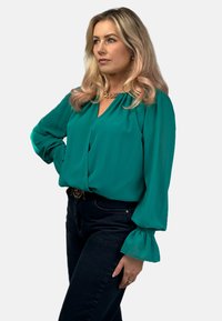 Turkusowa bluzka z długimi, bufiastymi rękawami, miękkiego materiału i dekoltem w kształcie litery V. W połączeniu z ciemnymi dżinsami i ozdobnym paskiem.