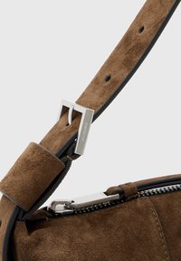 Bruine suede handtas met zwarte leren schouderriem, voorzien van een zilveren metalen gesp en ritshardware voor afsluiting. Gladde textuur en strak ontwerp.