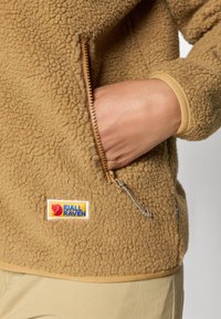 Giacca in pile color sabbia con superficie texturizzata, tasca con zip e patch con il logo Fjällräven. Presenta polsini elasticizzati e chiusura a zip.