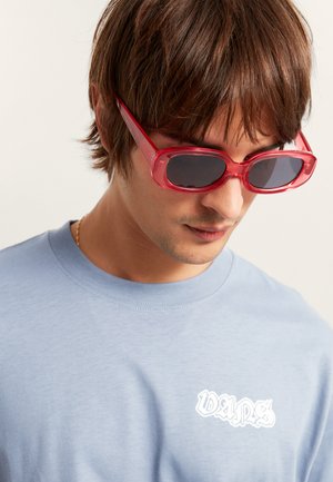 Jasnoniebieska bawełniana koszulka z białym logo. Przezroczyste czerwone okulary przeciwsłoneczne z ciemnymi soczewkami, elegancki design i zaokrąglone krawędzie.