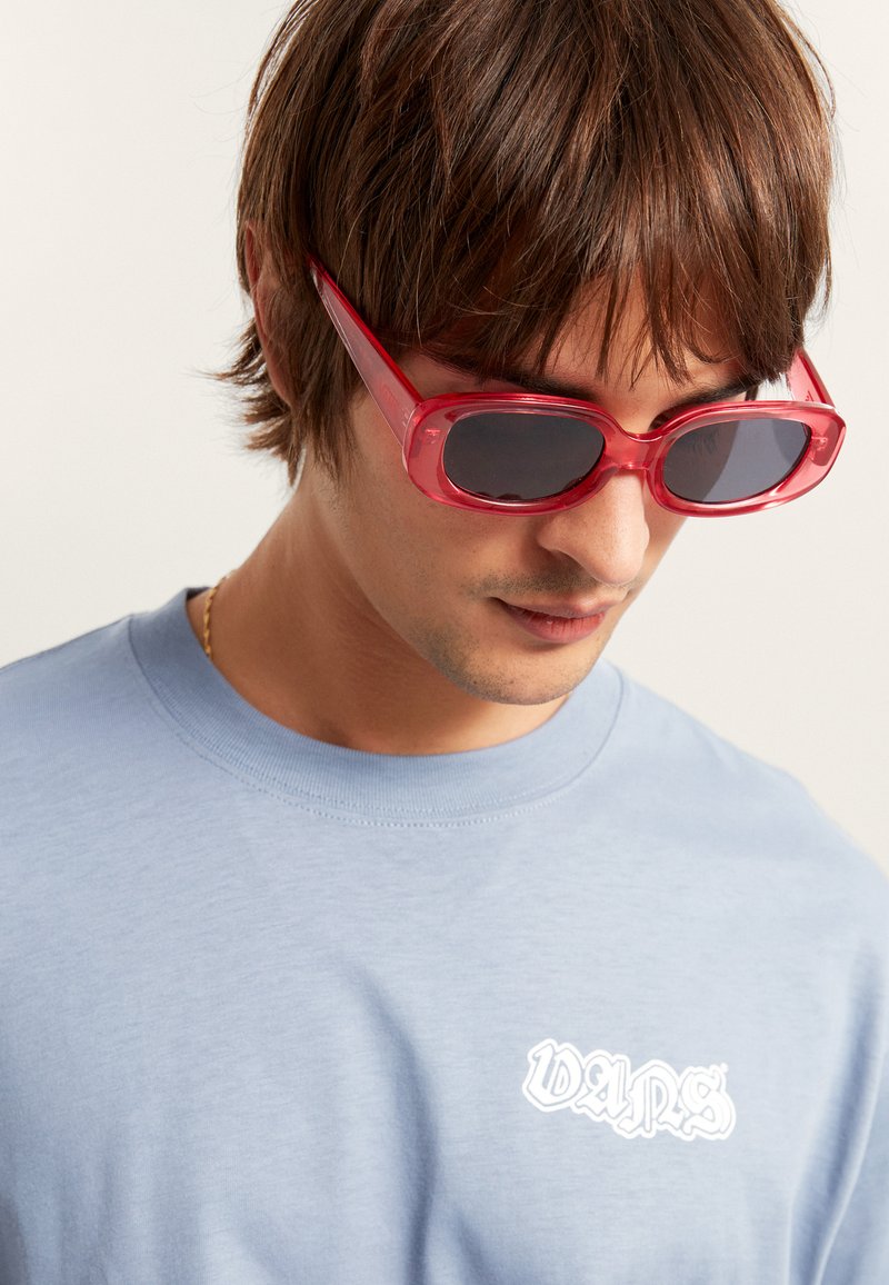 T-shirt en coton bleu clair avec un logo blanc. Lunettes de soleil rouges transparentes avec des verres foncés, au design élégant et aux bords arrondis.