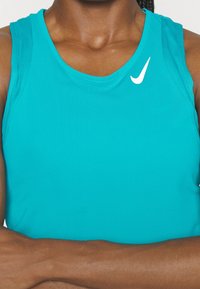 Personne portant un maillot de sport sans manches Nike turquoise avec un logo blanc, bras croisés sur la poitrine.