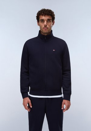 Uomo con capelli ricci indossa una giacca blu navy con zip e un piccolo logo rosso e bianco sul petto, in piedi davanti a uno sfondo chiaro e semplice.