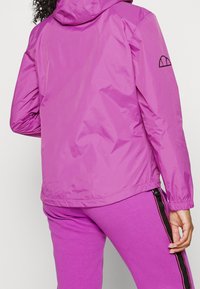 Person klädd i en knallrosa huvajacka och matchande byxor med svart och rosa sidostrip. Sedd bakifrån.