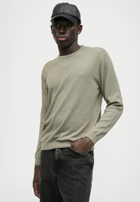 Maglione color khaki con texture a costine, scollatura rotonda e maniche lunghe, abbinato a jeans grigio scuro e un berretto di pelle nera.