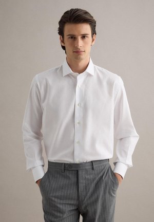 Giovane uomo in piedi con le mani nelle tasche, indossa una camicia bianca con bottoni e pantaloni grigi a righe sottili, su uno sfondo semplice.