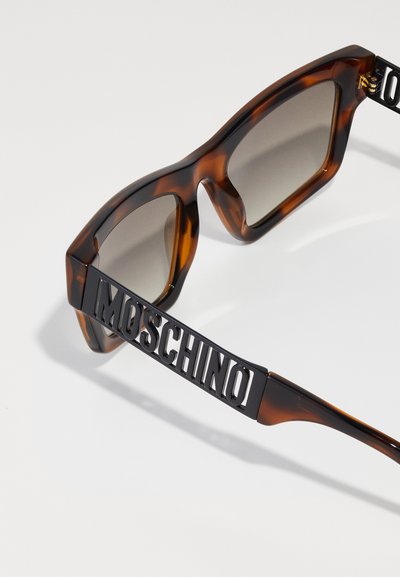 Ochelari de soare în carapace de țestoasă, cu o ramă geometrică, lentile întunecate și un accent îndrăzneț cu logo-ul „MOSCHINO” în negru pe laterale.