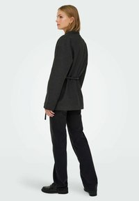 Graue Langarmjacke mit tailliertem Gürtel, kombiniert mit schwarzen Hosen und Schuhen. Abgerundeter Kragen und sichtbare Details aus strukturiertem Stoff.
