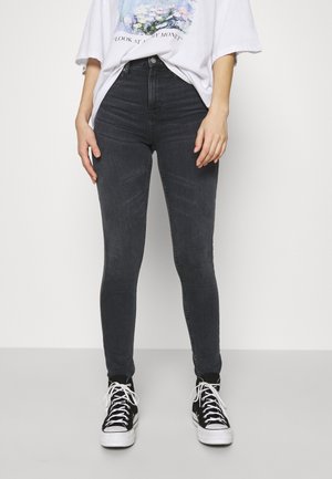 Personne portant un jean skinny gris foncé taille haute, un t-shirt blanc à motif graphique, et des baskets montantes noires avec des lacets blancs.