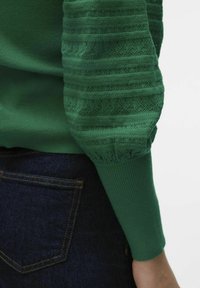 Zeleni pleteni pulover z teksturiranimi rokavi in diamantnimi vzorci, rebrastimi manšetami in gladkim telesom. Uporabljen z temnimi kavbojkami.