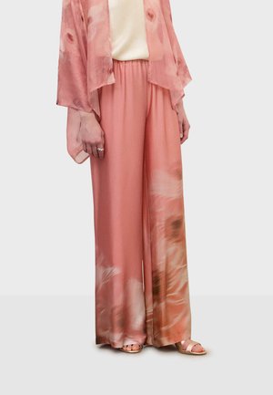 Kvinde iført lyserøde vidde-ben bukser med tie-dye, matchende gennemsigtig cardigan, cremefarvet top og sandaler i rosaguld, stående mod en ensfarvet baggrund.