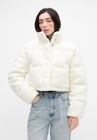 Krátka biela chlpová bunda s dvoma bočnými vreckami, vysokým golierom a cvokovými uzávermi, v kombinácii s džínsami svetlomodrej farby s vysokým pásom.