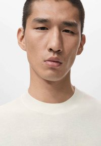 Světlý krémový svetr s žebrovaným kulatým výstřihem. Tkanina je texturovaná a přiléhavá. Pozadí je neutrální a nenápadné.